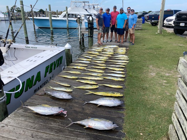 A small group of people stand on the boardwalk next to their boat and display two dozen fish they caught.