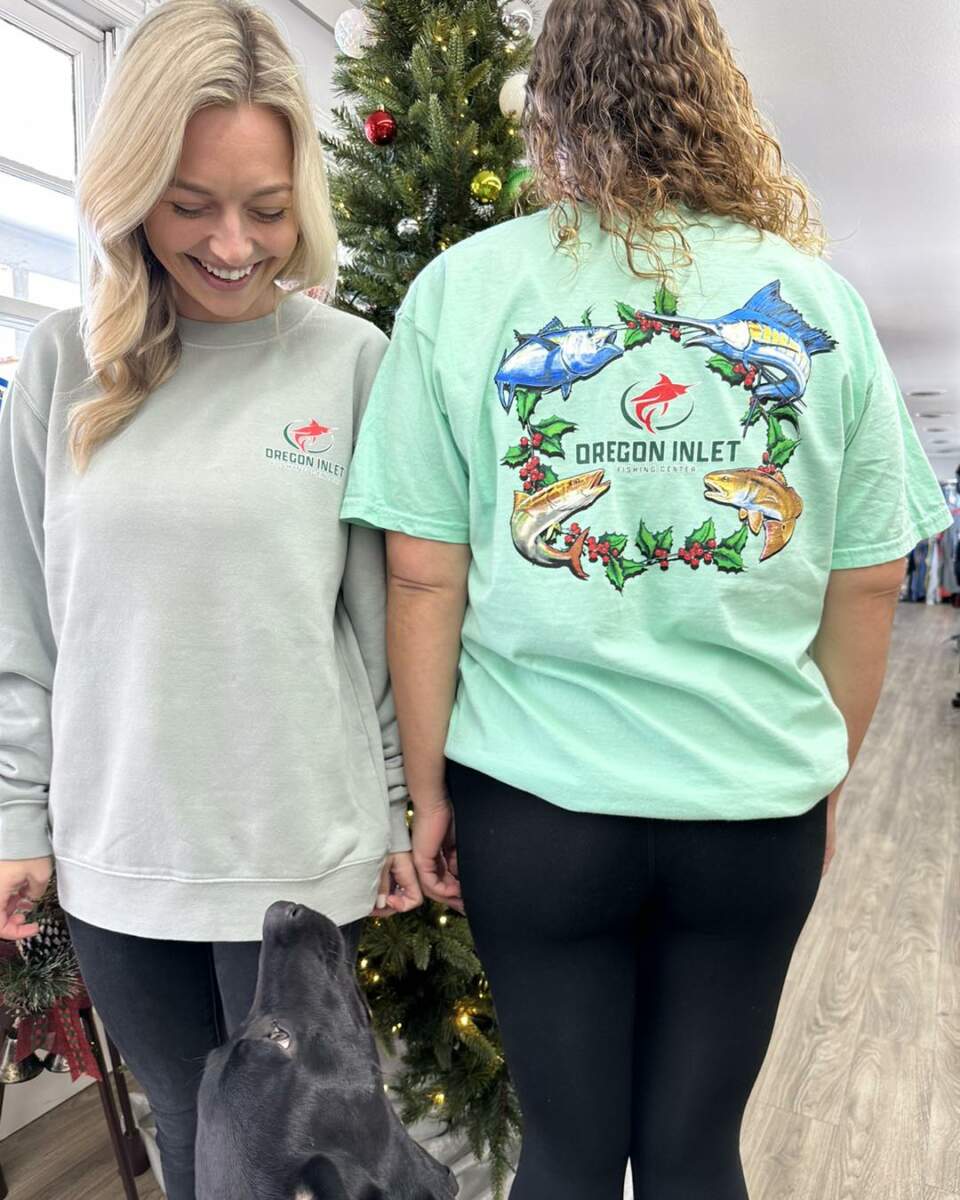 Two people and a dog model Oregon Inlet Fishing Center apparel in front of a Christmas tree.