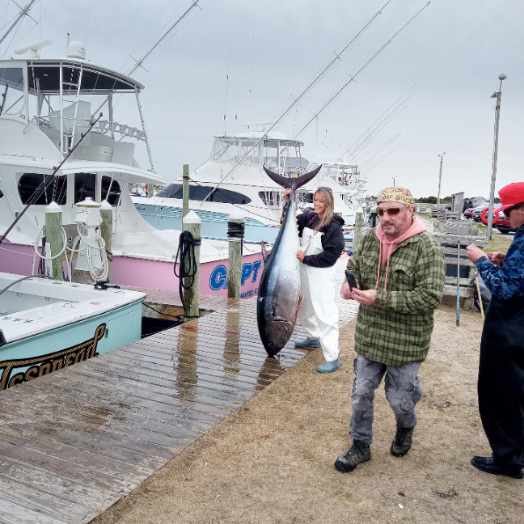 A person in white waders stands on the dock and holds up a large tuna they caught.