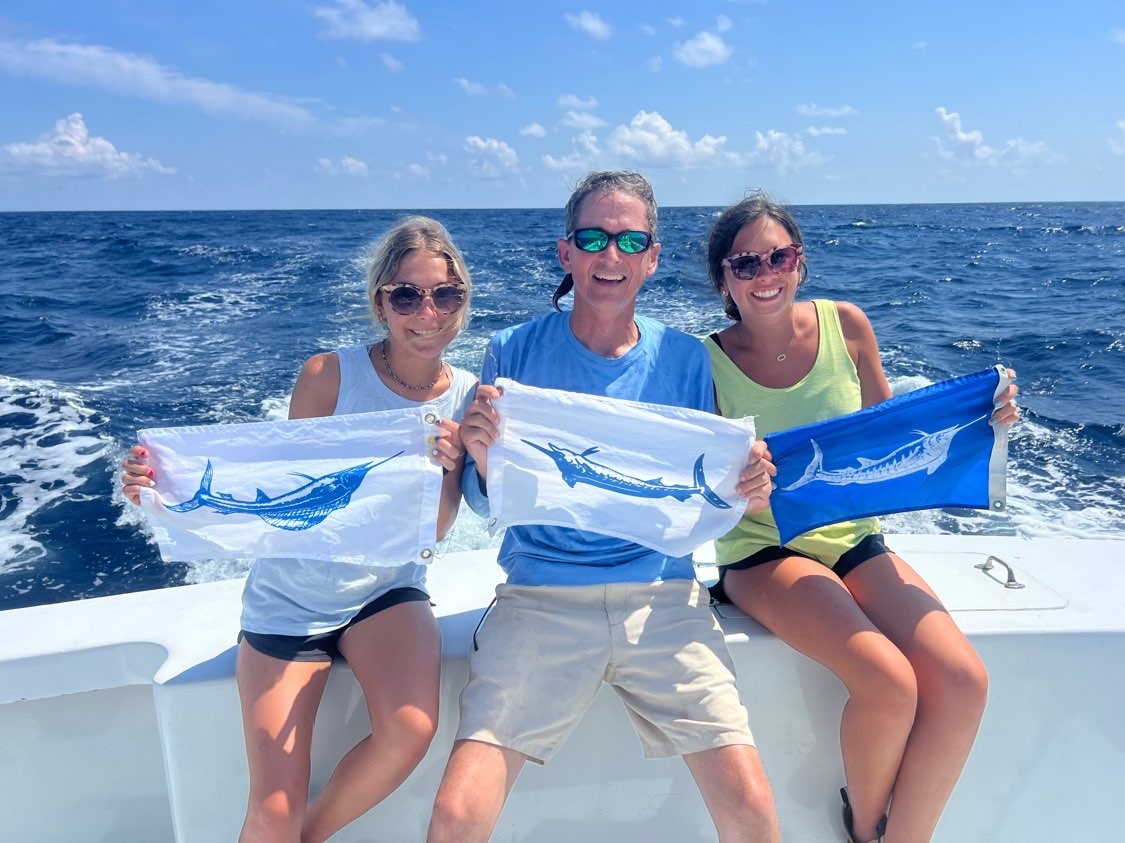 Three people hold up flags with fish on them as they ride on the back of a fishing charter in Outer Banks, NC.